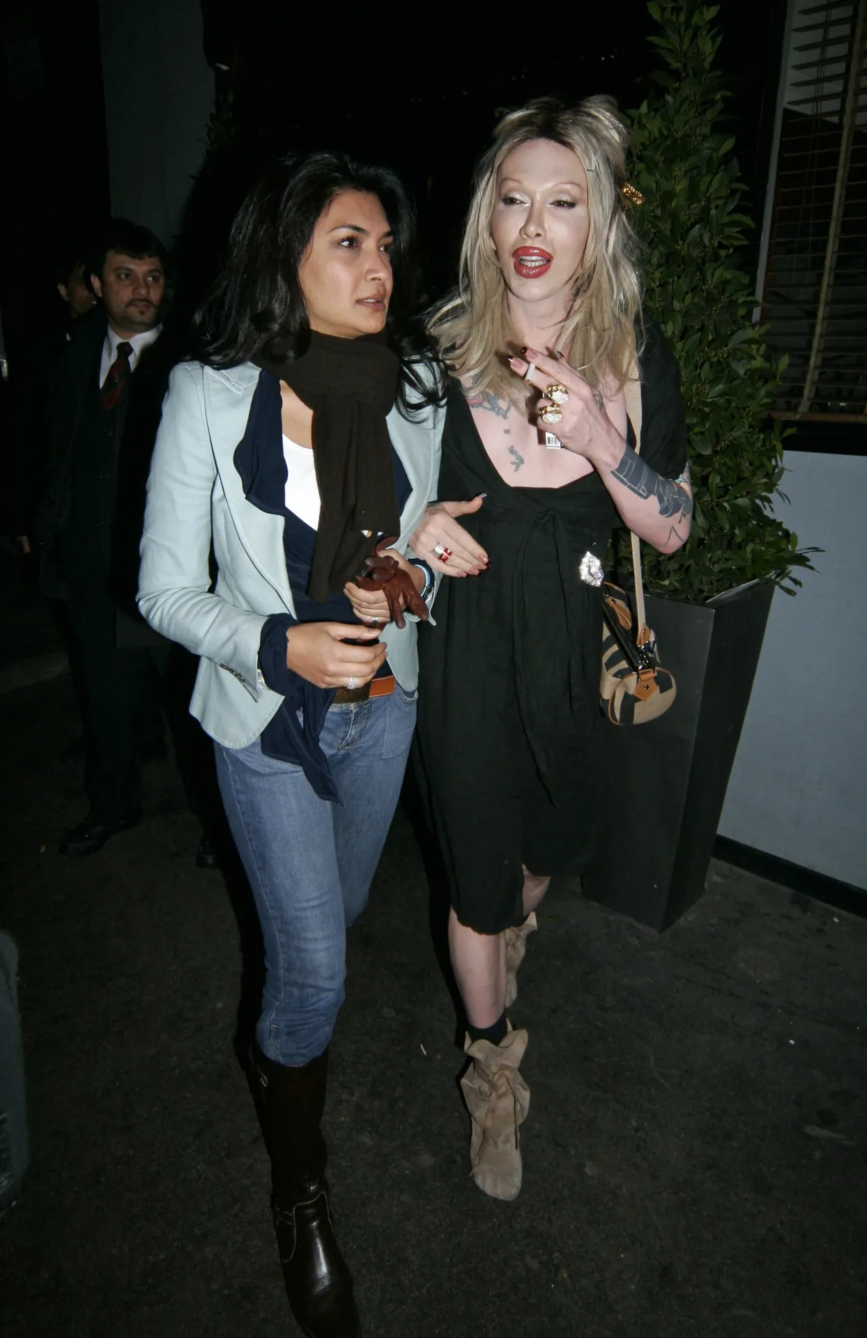 Pete Burns of Dead or Alive, with long blonde hair and a black dress, walking beside Faria Alam in a pale blue jacket outside the Groucho Club in Soho at night, February 2006.