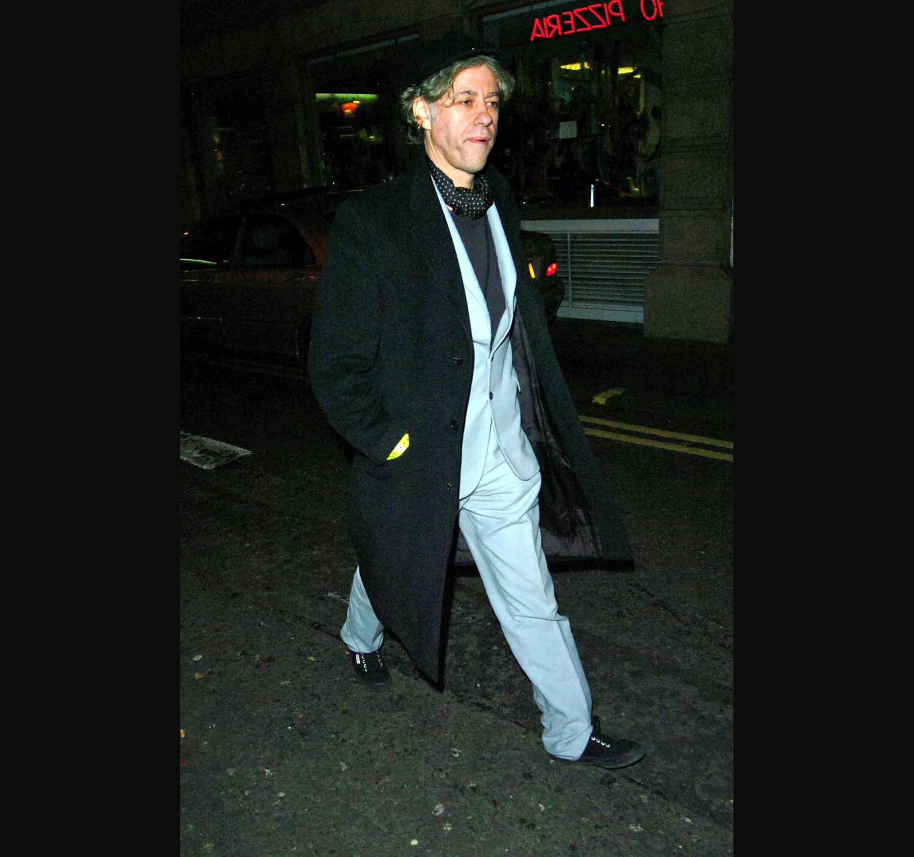 Bob Geldof, in a long black coat and pale suit, walks off down the street.