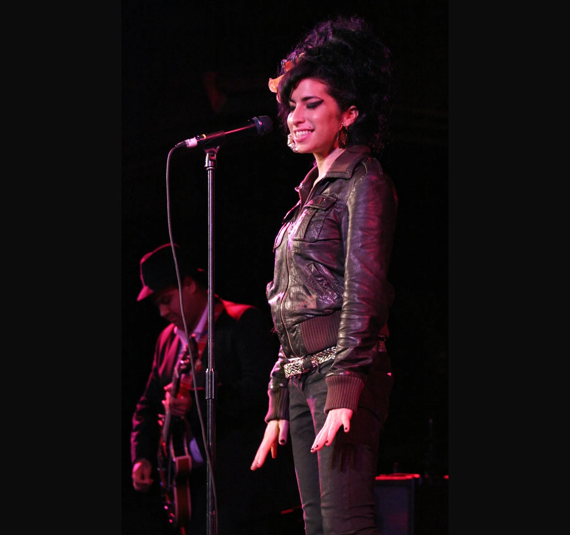 In a dark leather jacket, smiling as she sings, with a musician in a hat just visible behind her.