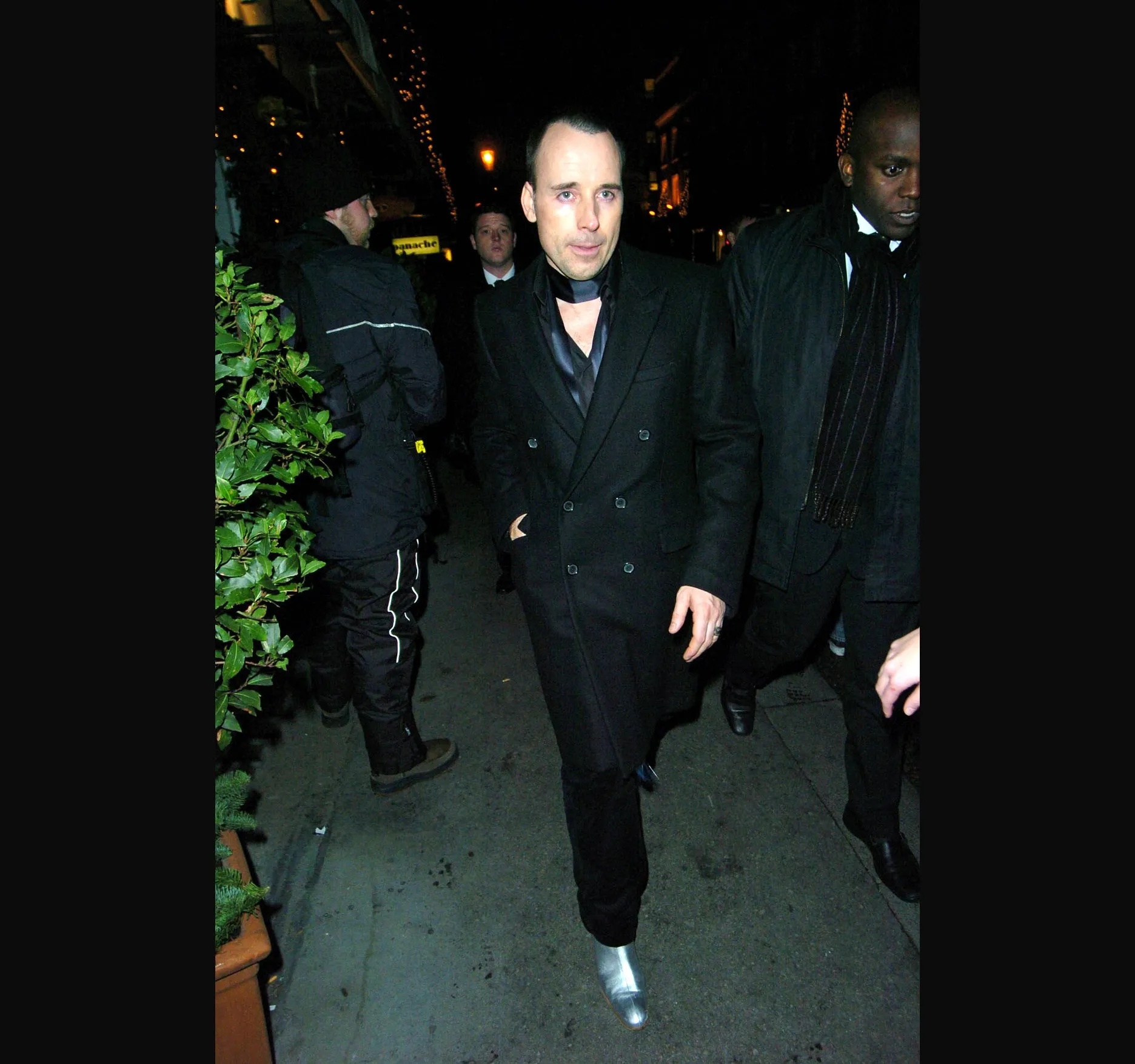 DAVID FURNISH. David Furnish walks along the pavement in a black double breasted coat, with a black scarf at his neck and silver shoes.