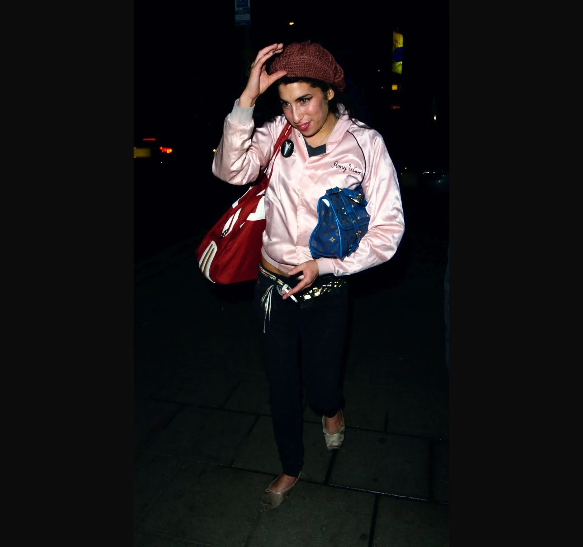 Amy Winehouse lifting her pink beret with one hand as she walks along a pavement at night, a red bag on her shoulder.