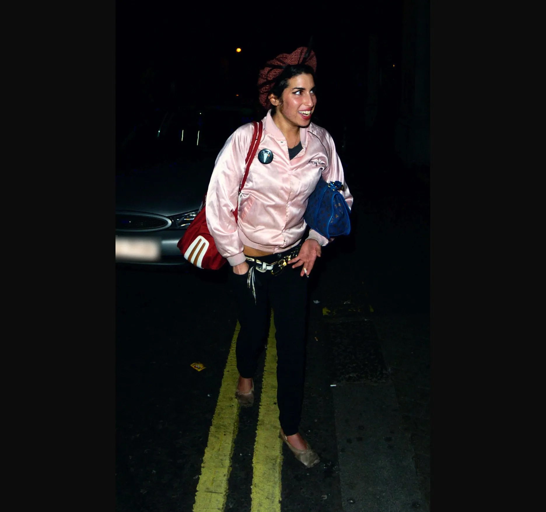 Amy Winehouse smiling in a pink satin jacket and beret beside double yellow lines and a parked car at night.