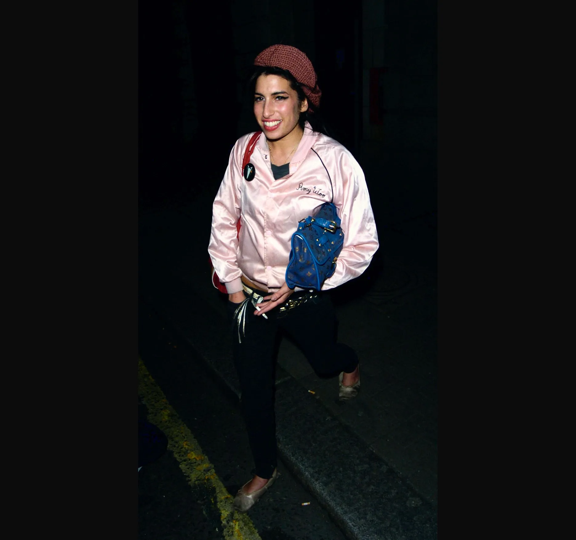 Amy Winehouse walking towards the camera on a dark street, smiling, in a pink satin jacket and black trousers.