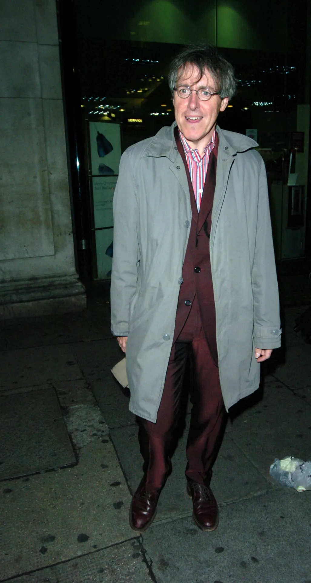 Griff Rhys Jones standing straight-faced in a grey raincoat and burgundy suit outside a West End bar at night.
