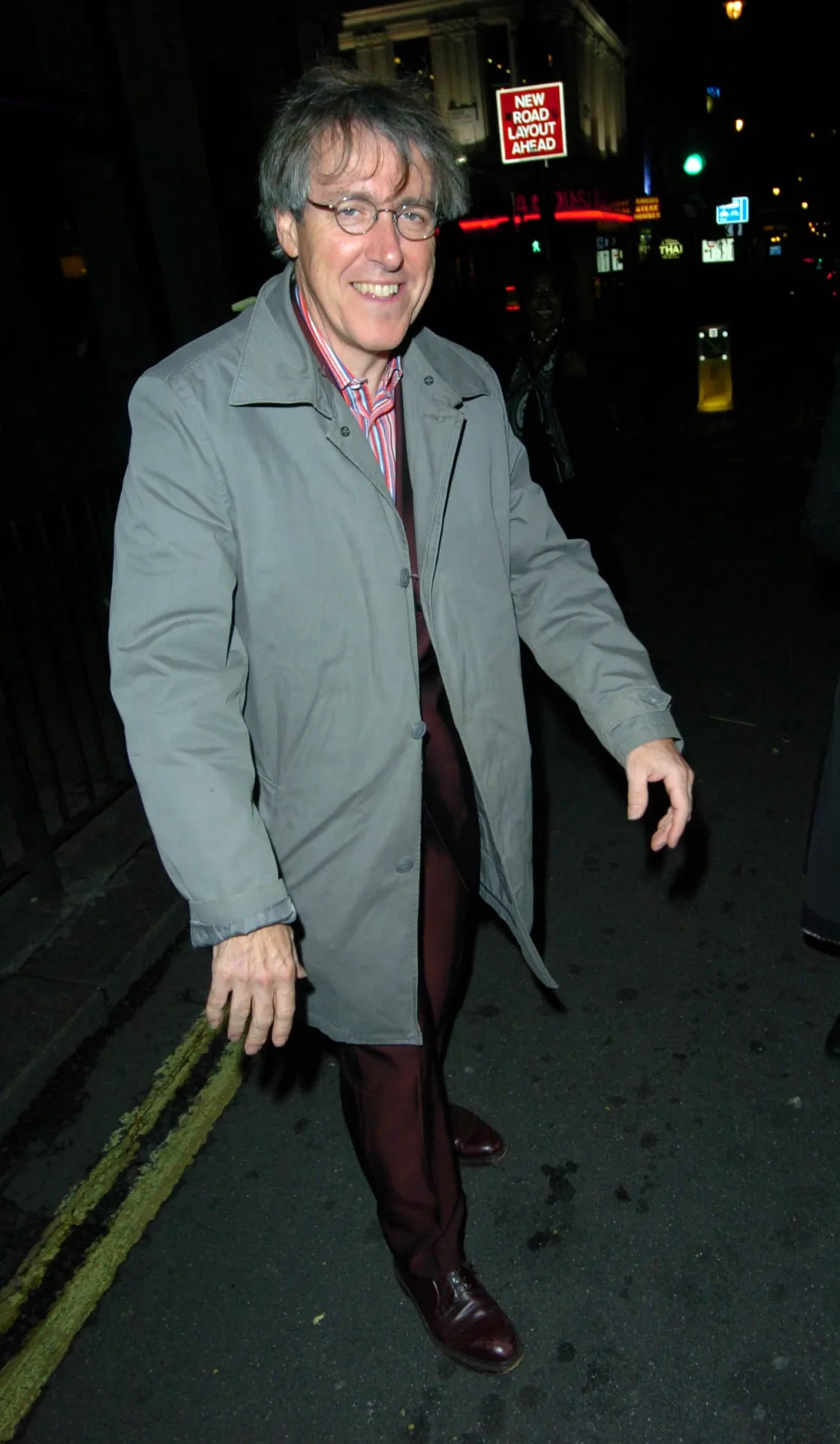 Griff Rhys Jones walking towards the camera with his arms held out from his sides, smiling, on a London street at night.