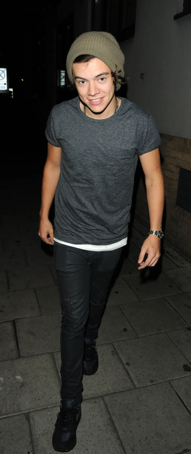 Harry Styles smiling, in a khaki beanie, grey T-shirt and black jeans, walking along a London pavement at night.