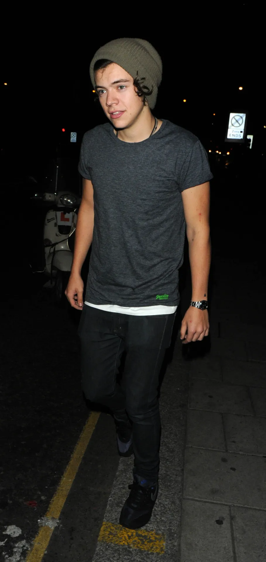Harry Styles in a beanie and grey T-shirt walking towards the camera beside a yellow kerb line.