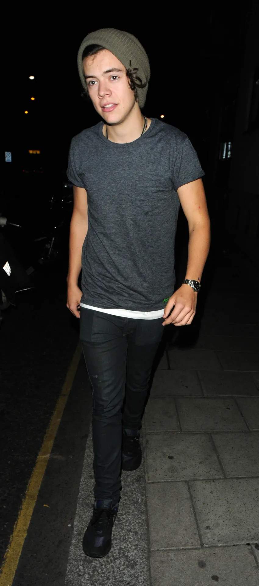 Harry Styles in a beanie, grey T-shirt and black jeans striding along a dark street.