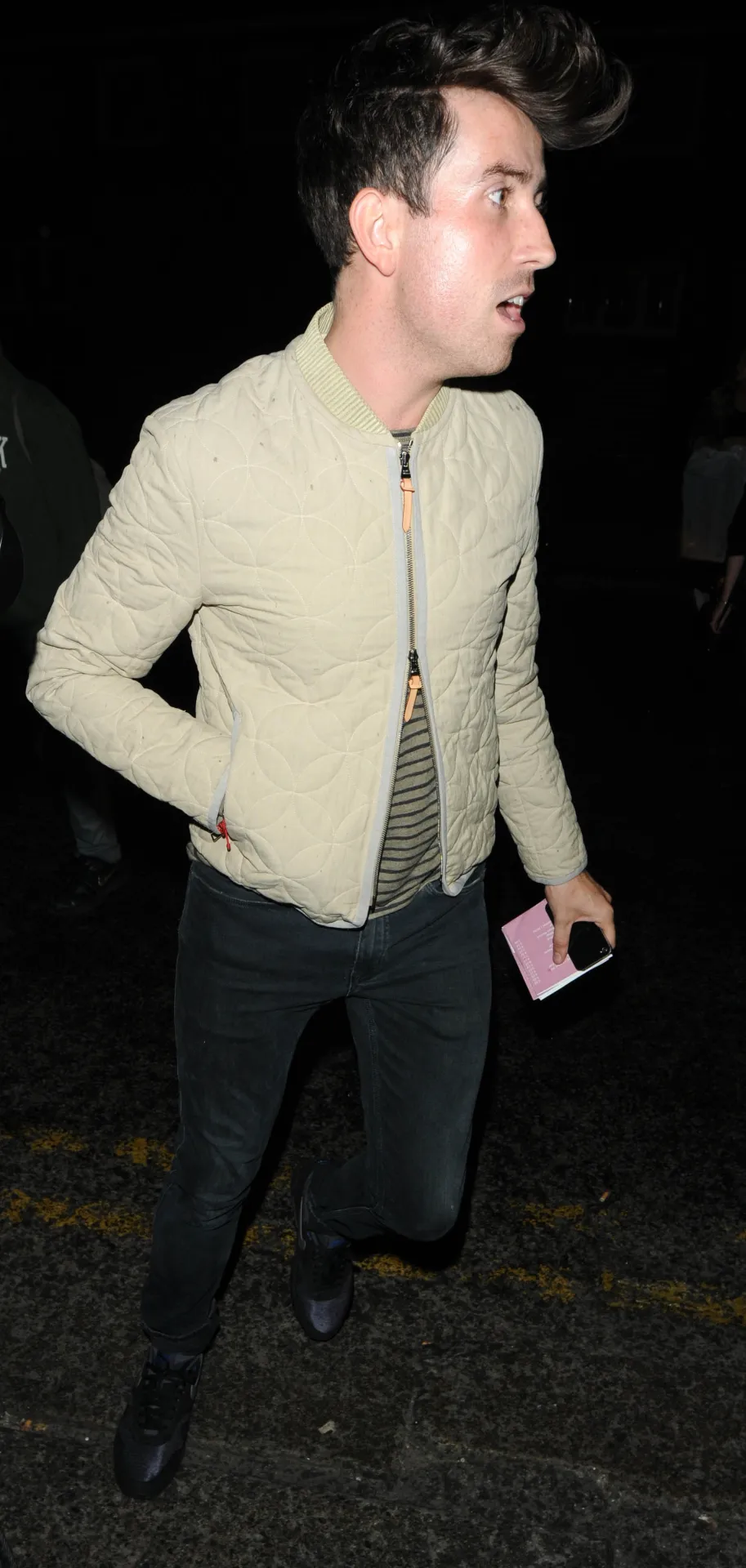 Nick Grimshaw with a quiff, in a cream zip jacket and striped top, holding a pink phone at night.