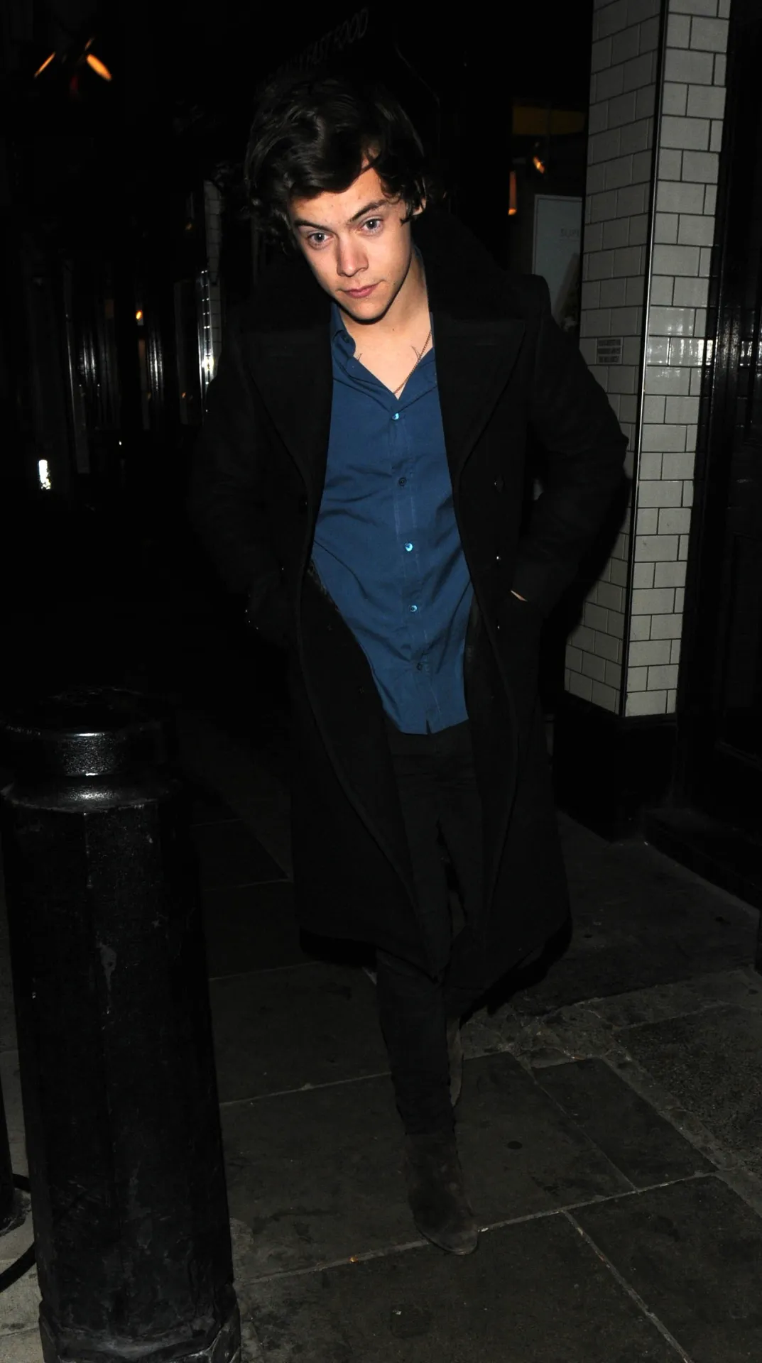 Harry Styles with longer hair, in a long black coat and blue shirt, walking past a white tiled wall at night.