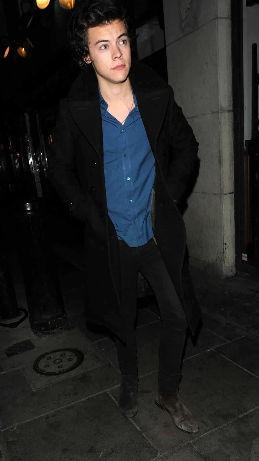 Harry Styles in a long black coat and open blue shirt, hands in pockets, on a Soho pavement.