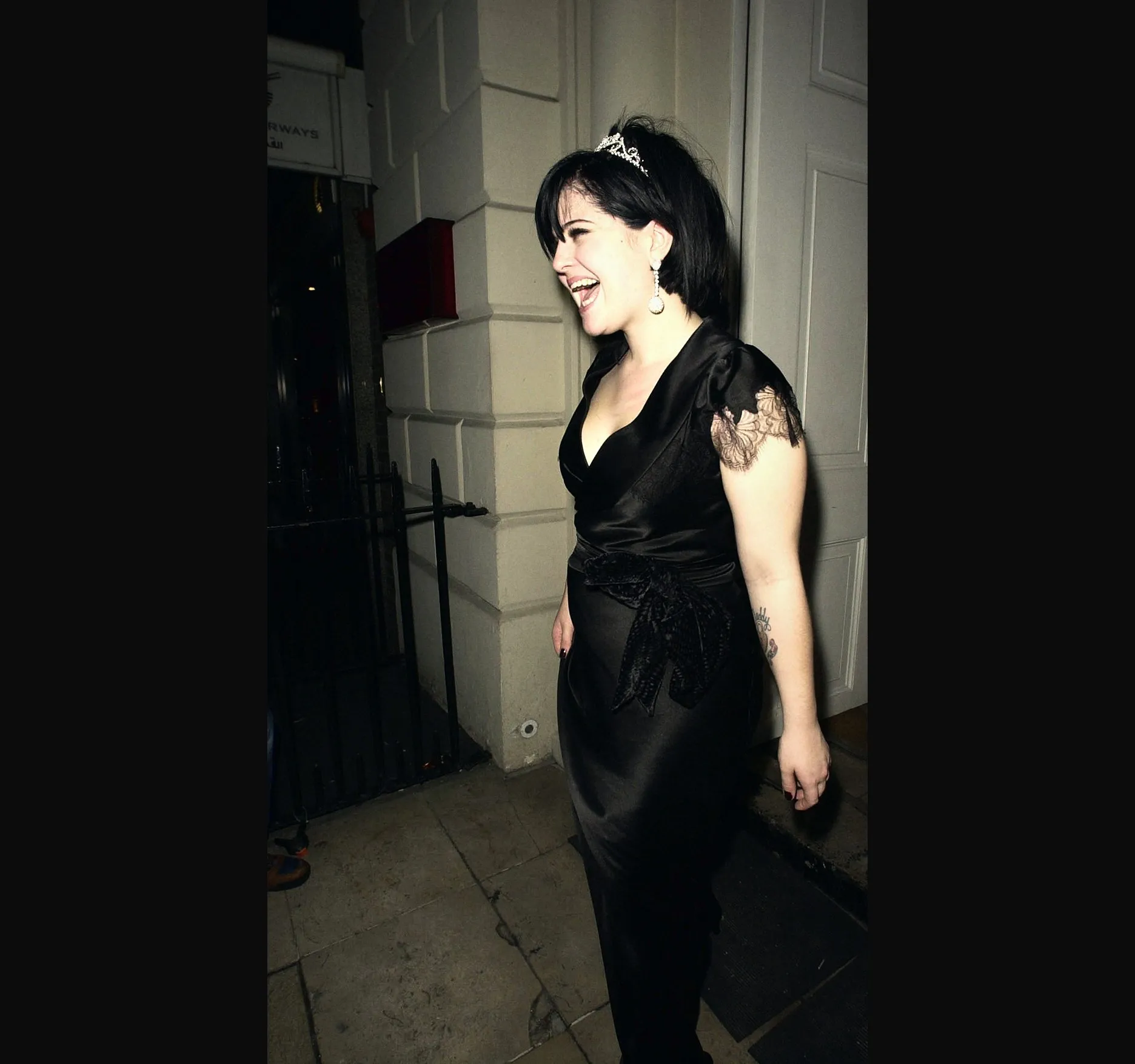 Kelly Osbourne laughing in a long black satin gown and a sparkly tiara, standing by the white columned doorway of Sketch at n