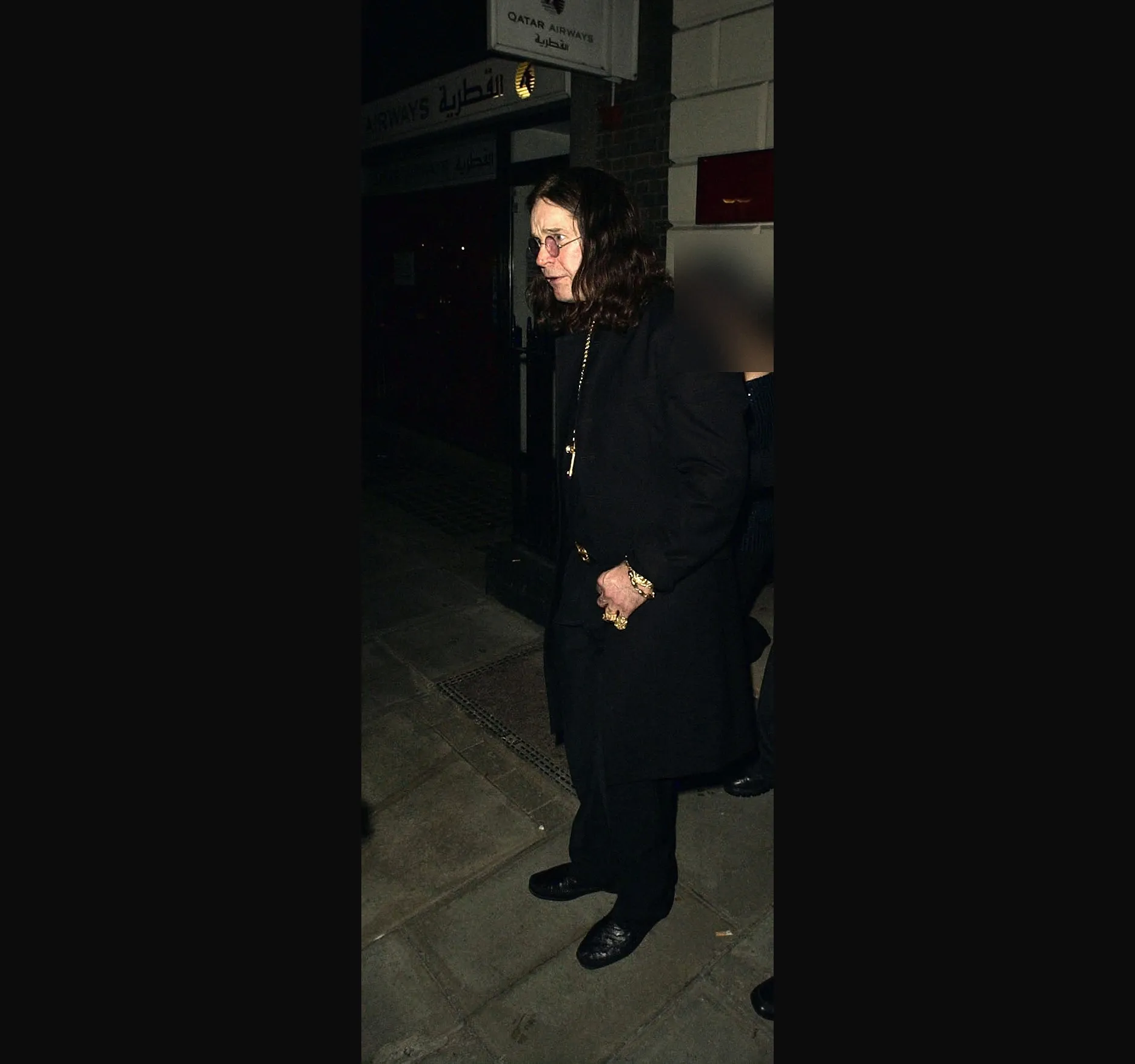 Ozzy Osbourne with long dark hair and round glasses, in a long black coat with a gold cross, standing on a Mayfair pavement a