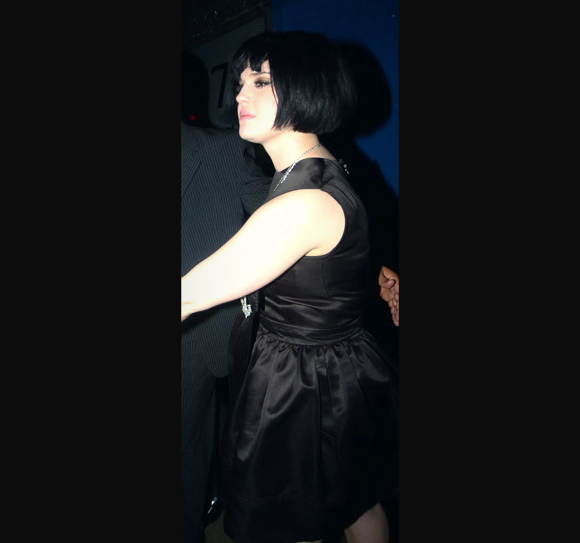 Kelly Osbourne in profile with a black bob and a black satin sleeveless dress, one arm outstretched, outside a Soho club at n