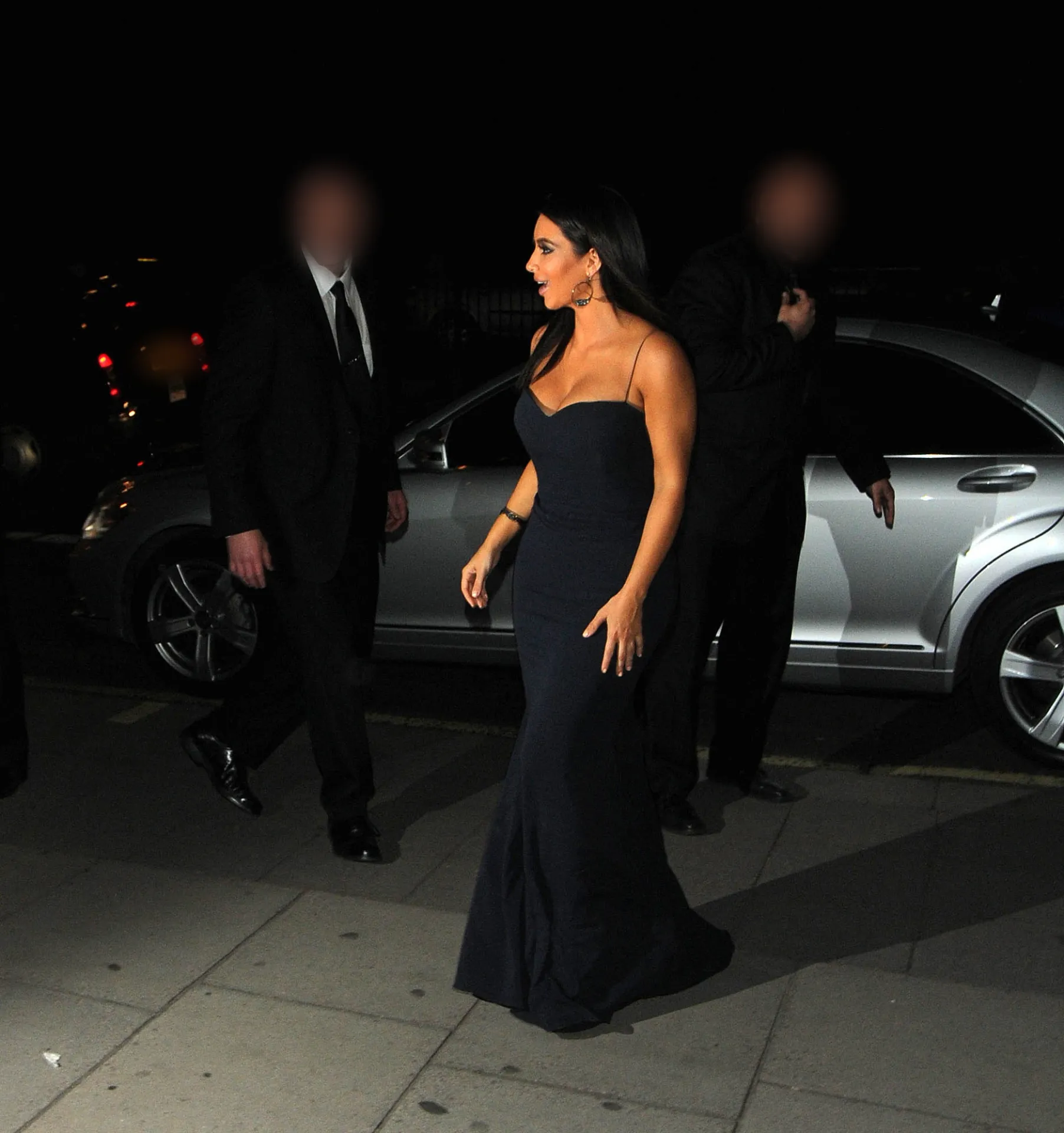 Kim Kardashian in a long navy strapless gown walking past a silver car outside the Athenaeum Hotel at night.