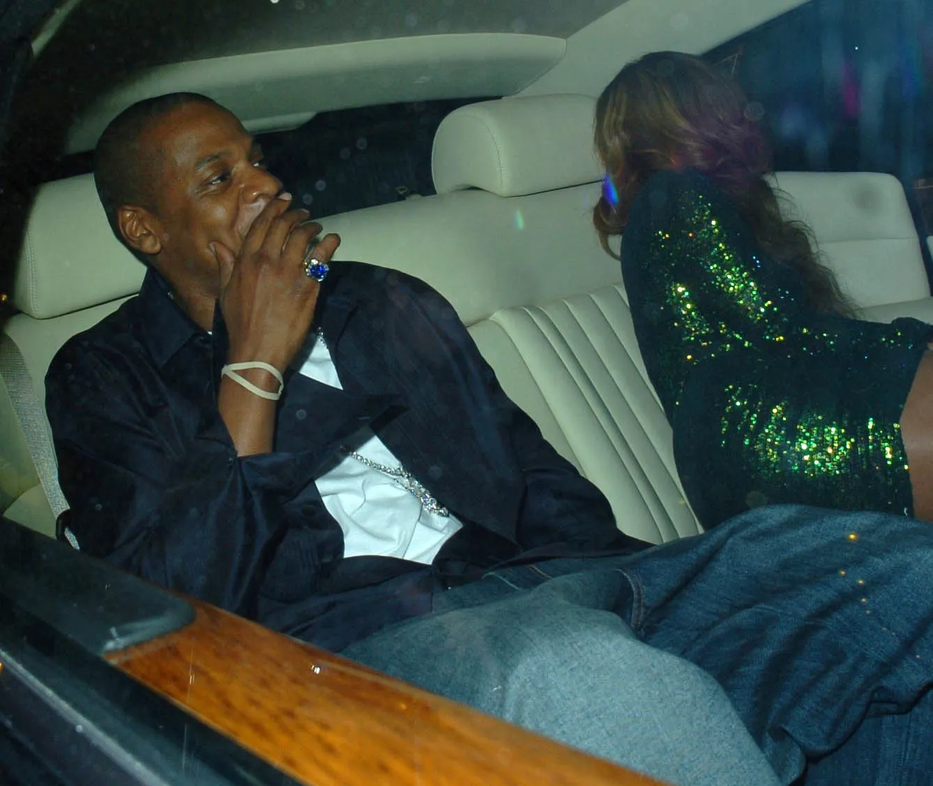 Jay-Z, in a dark shirt with a hand raised to his face, sits in the back of a car with cream leather seats beside Beyoncé in a