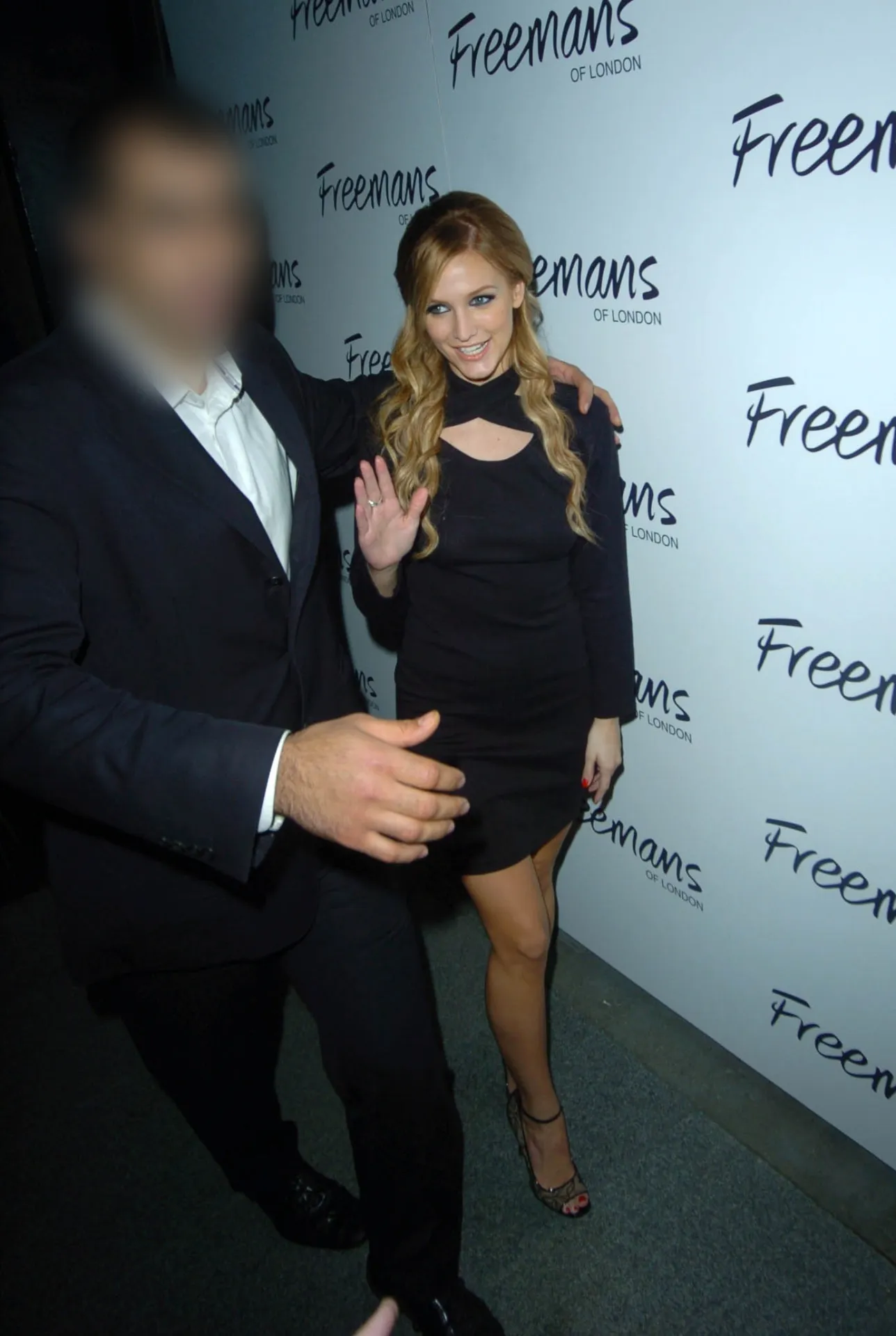 Ashlee Simpson in a short black dress waves in front of a white branded backdrop at a party.