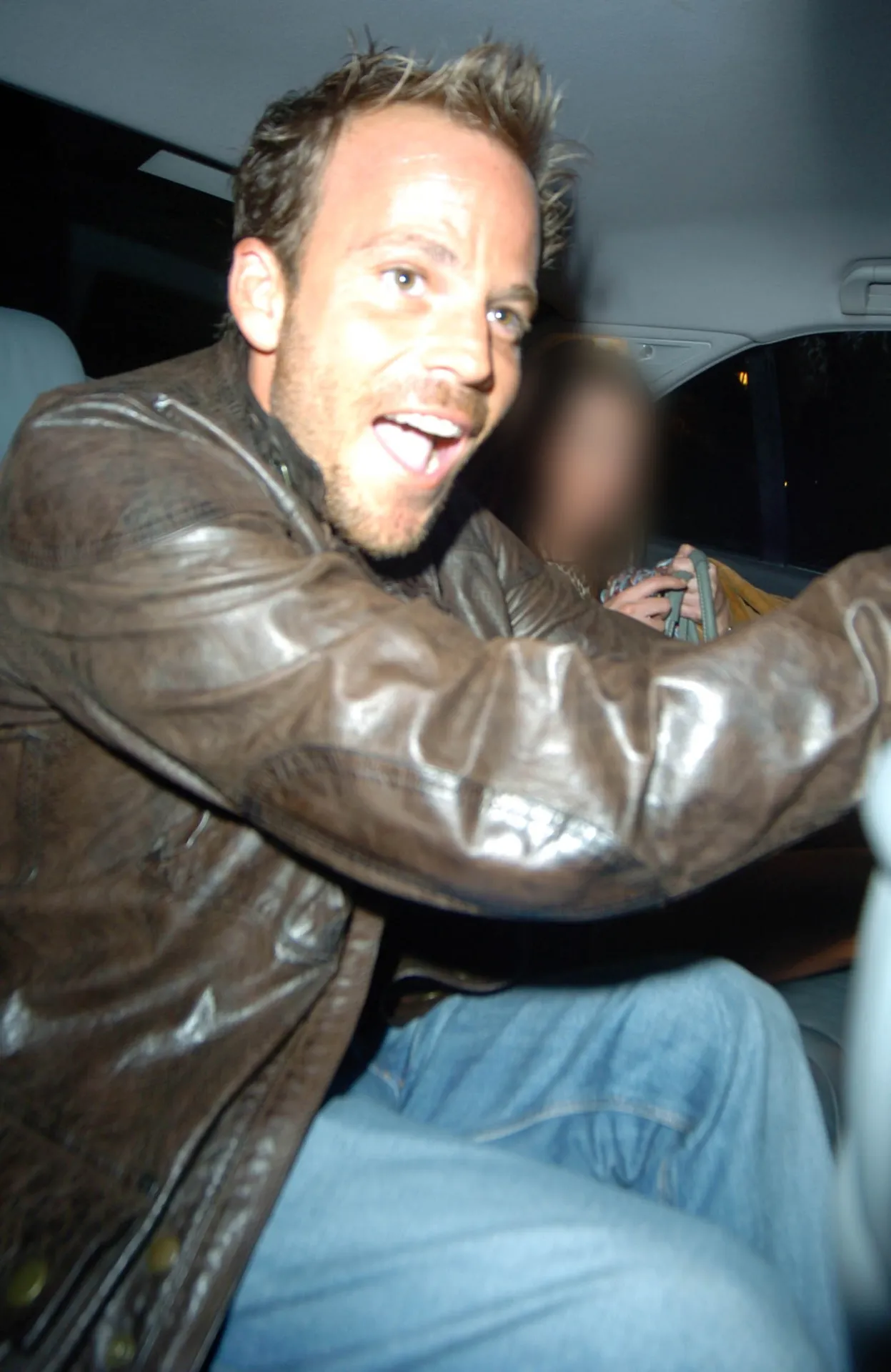 Stephen Dorff in a brown leather jacket grins at the camera from the back seat of a car at night.