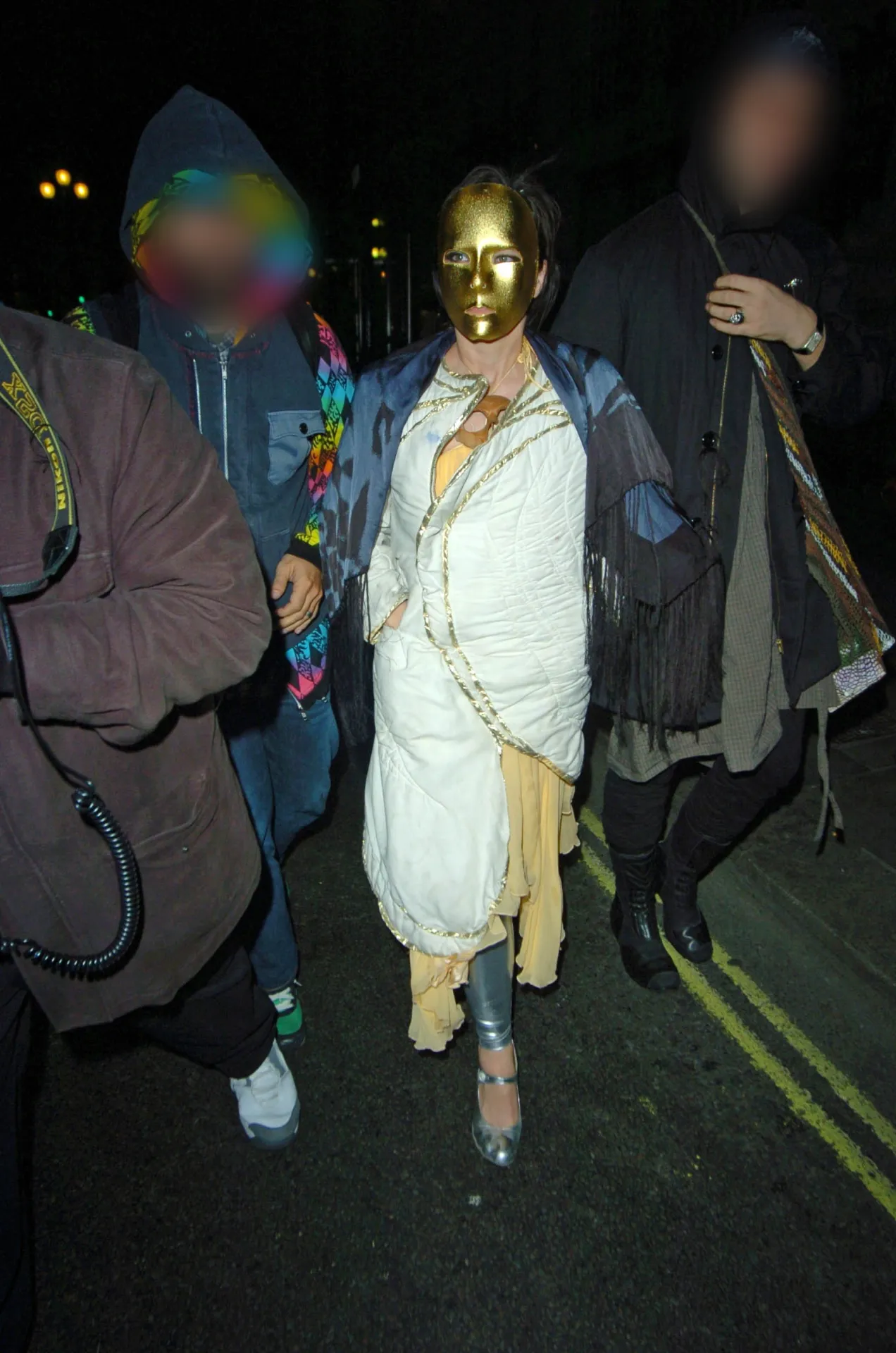 Björk in a gold face mask and a white quilted wrap walks along a London street at night with hooded companions.