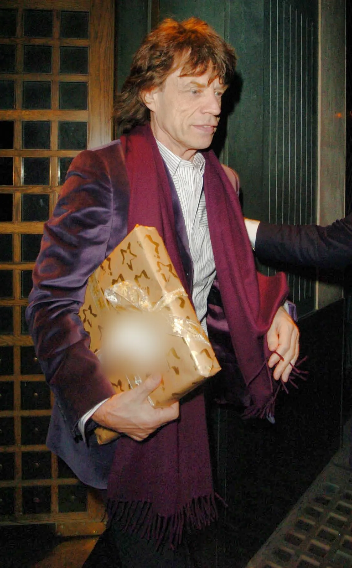 Mick Jagger in a purple velvet jacket and burgundy scarf leaving The Ivy restaurant at night, holding a gift wrapped in gold