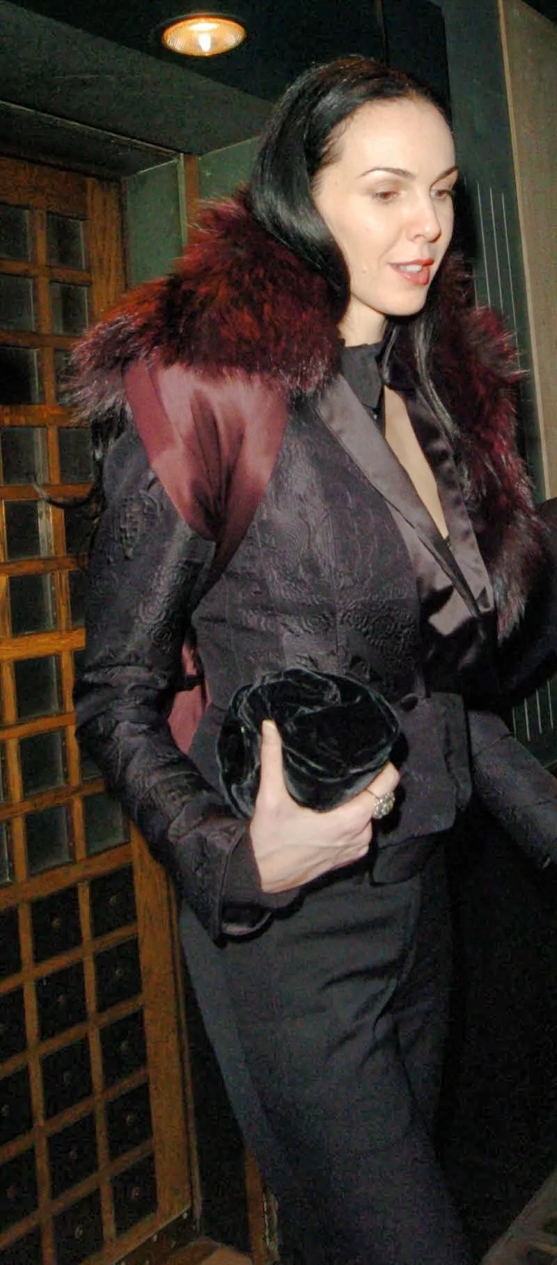 L'Wren Scott in a dark patterned jacket with a deep red fur collar, holding a black velvet clutch, leaving The Ivy at night.