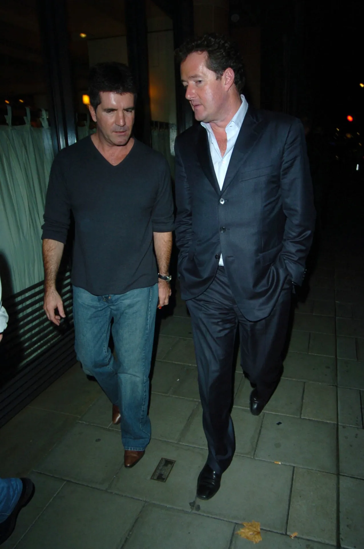 Simon Cowell in a dark V-neck top and jeans walks beside Piers Morgan in a navy suit, who turns to talk to him, on a Mayfair