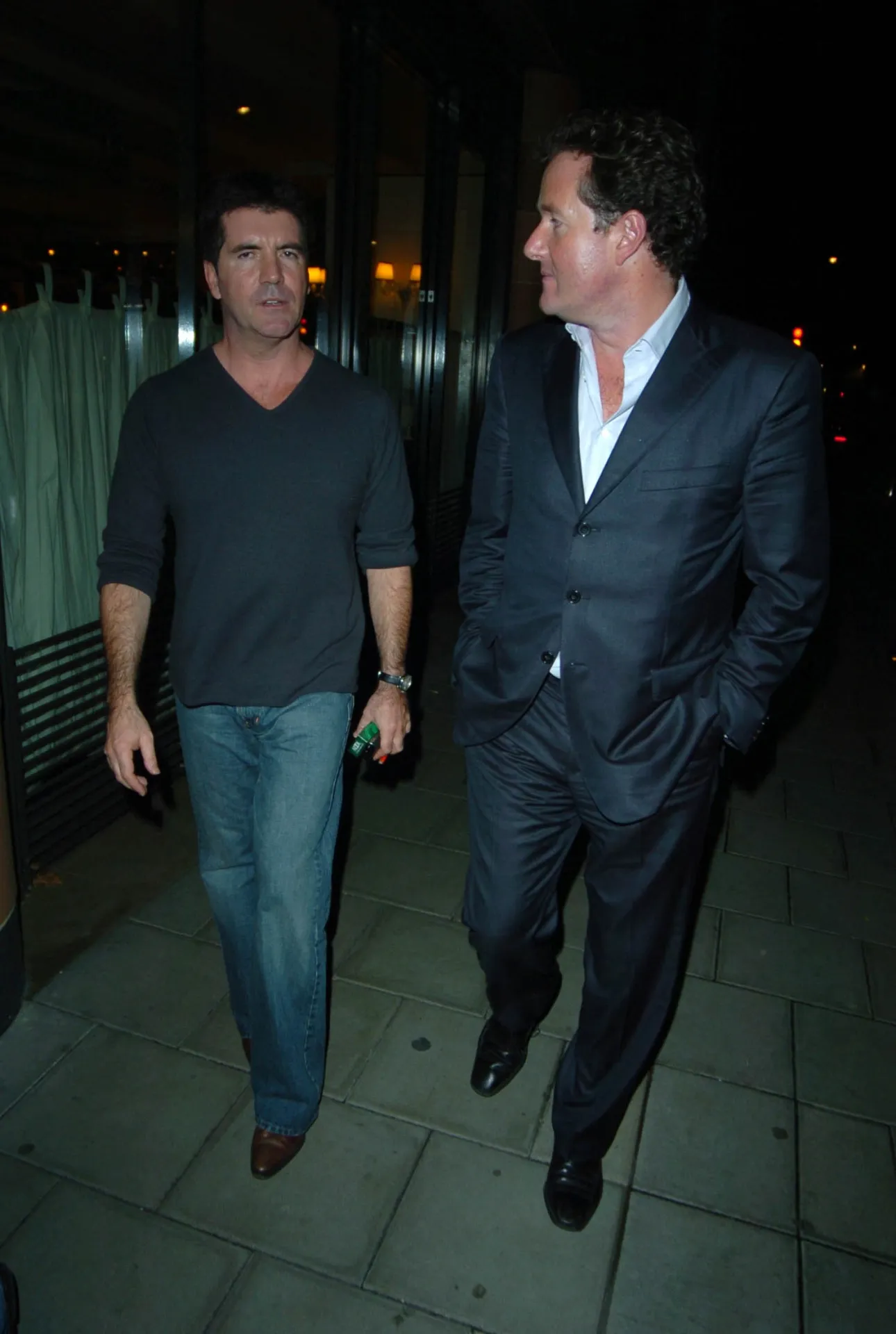 Simon Cowell and Piers Morgan walk side by side along a dimly lit Mayfair pavement, Morgan with one hand in his trouser pocke