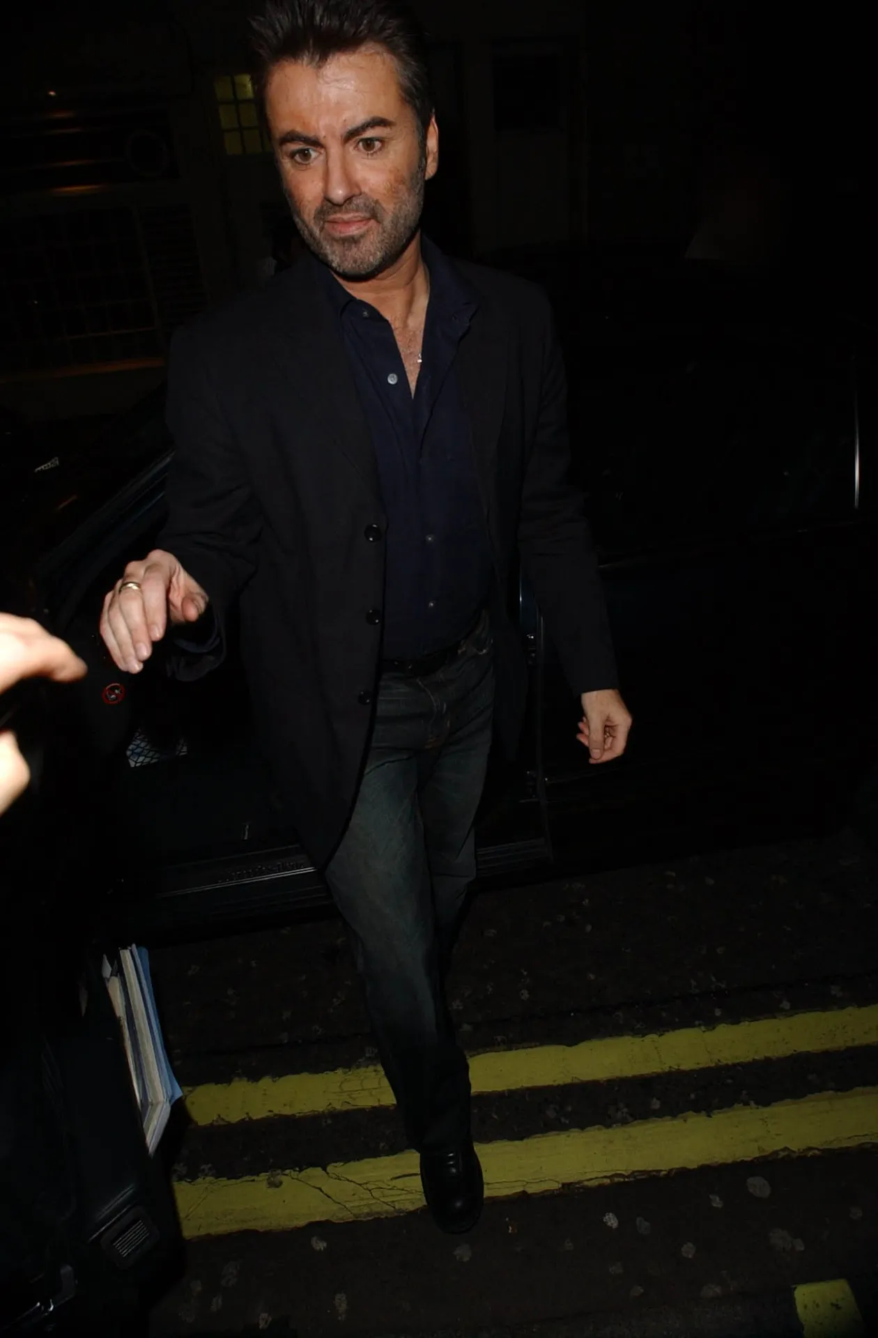 George Michael in a dark jacket and navy shirt stepping out of a car onto a pavement with double yellow lines at night.