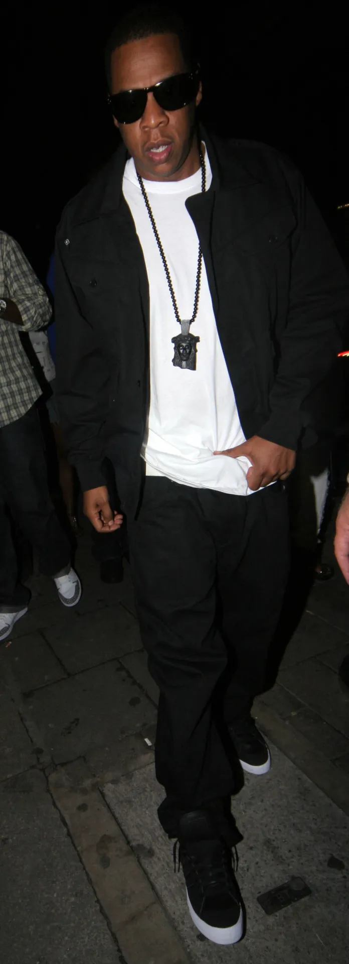 Jay-Z in a black jacket, white T-shirt and chain walking along a London pavement at night.