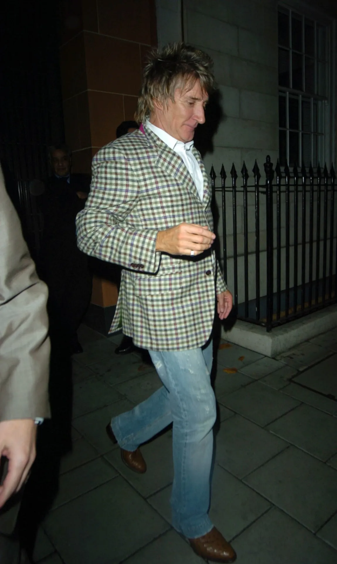 Rod Stewart in a check jacket and jeans walking along a Mayfair street at night.
