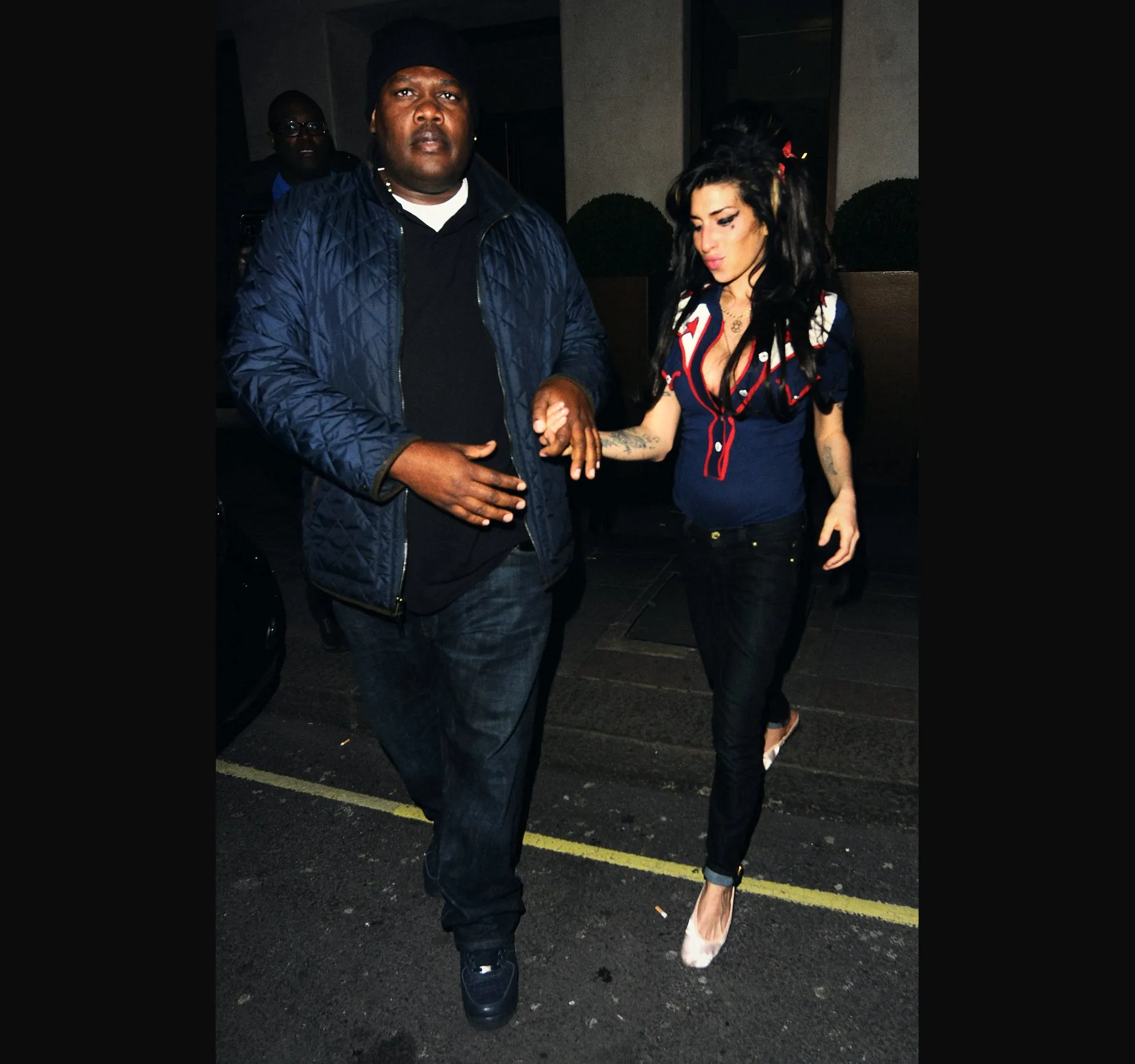 THE MAY FAIR, APRIL 2010. Amy Winehouse leaves The May Fair Hotel in Mayfair in the early hours of 16 April 2010, after a night out on 15 April.

She wears a navy top with red trim and dark jeans, with a man in a dark quilted jacket at her side.