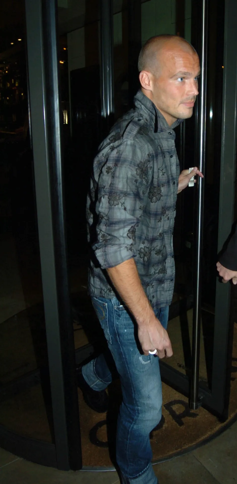 Freddie Ljungberg in a grey check shirt and jeans walking out through the revolving door of Cipriani in Mayfair at night.