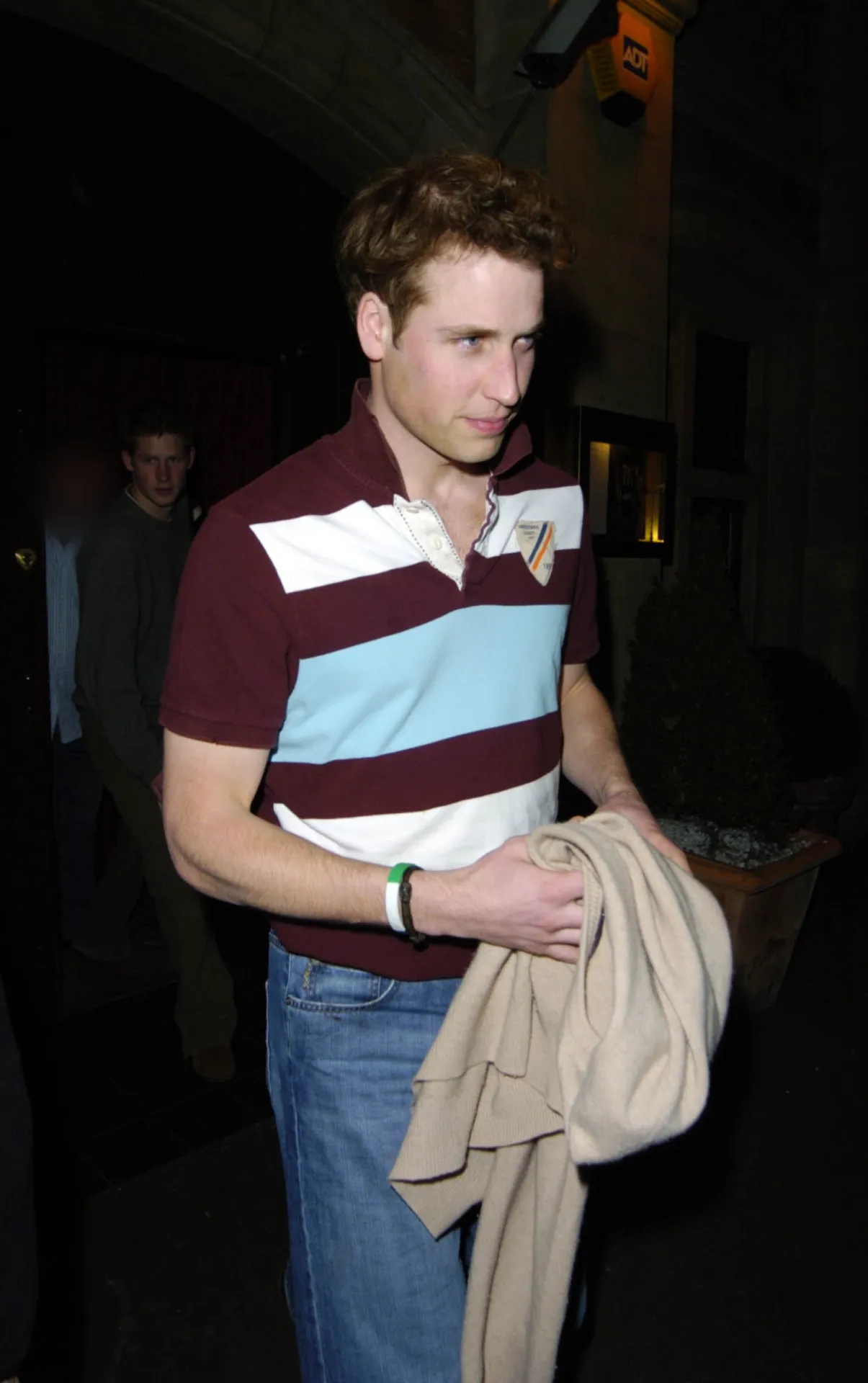 Prince William in a maroon, white and pale blue striped rugby shirt and jeans, a beige jacket over his arm, with Prince Harry