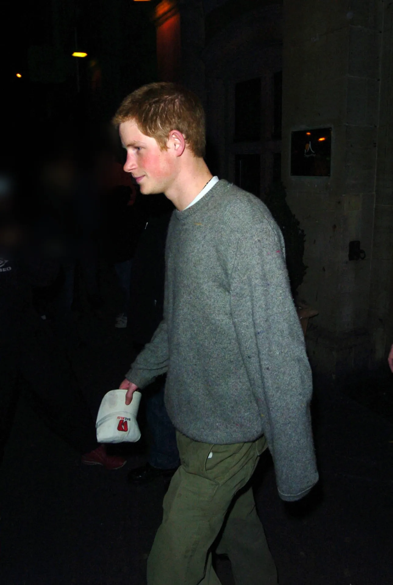 Prince Harry with short ginger hair in a grey jumper and khaki trousers, walking along a dark street at night holding a small