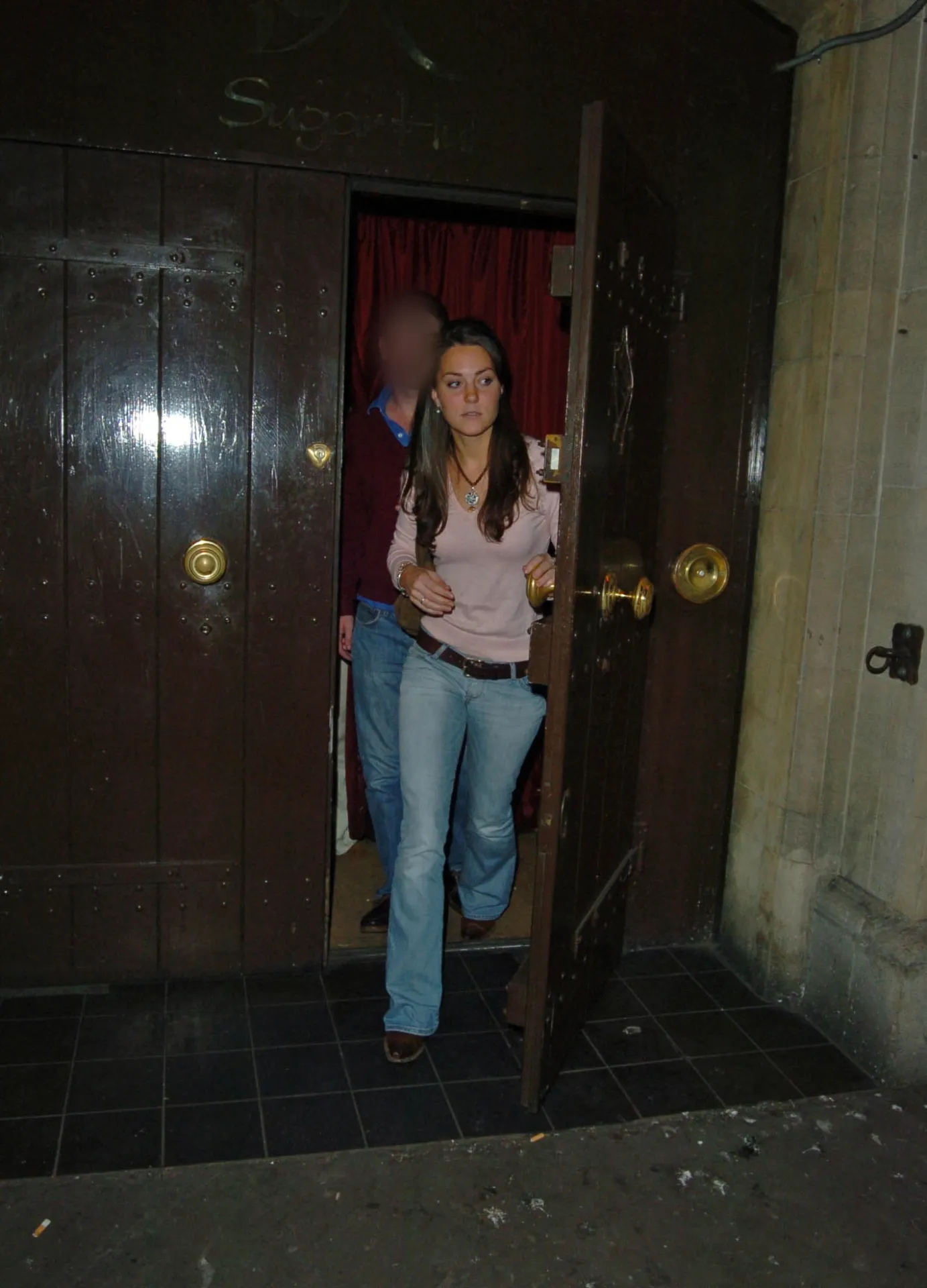 Kate Middleton with long dark hair in a pale pink top and flared blue jeans, stepping out of a large dark wooden doorway with