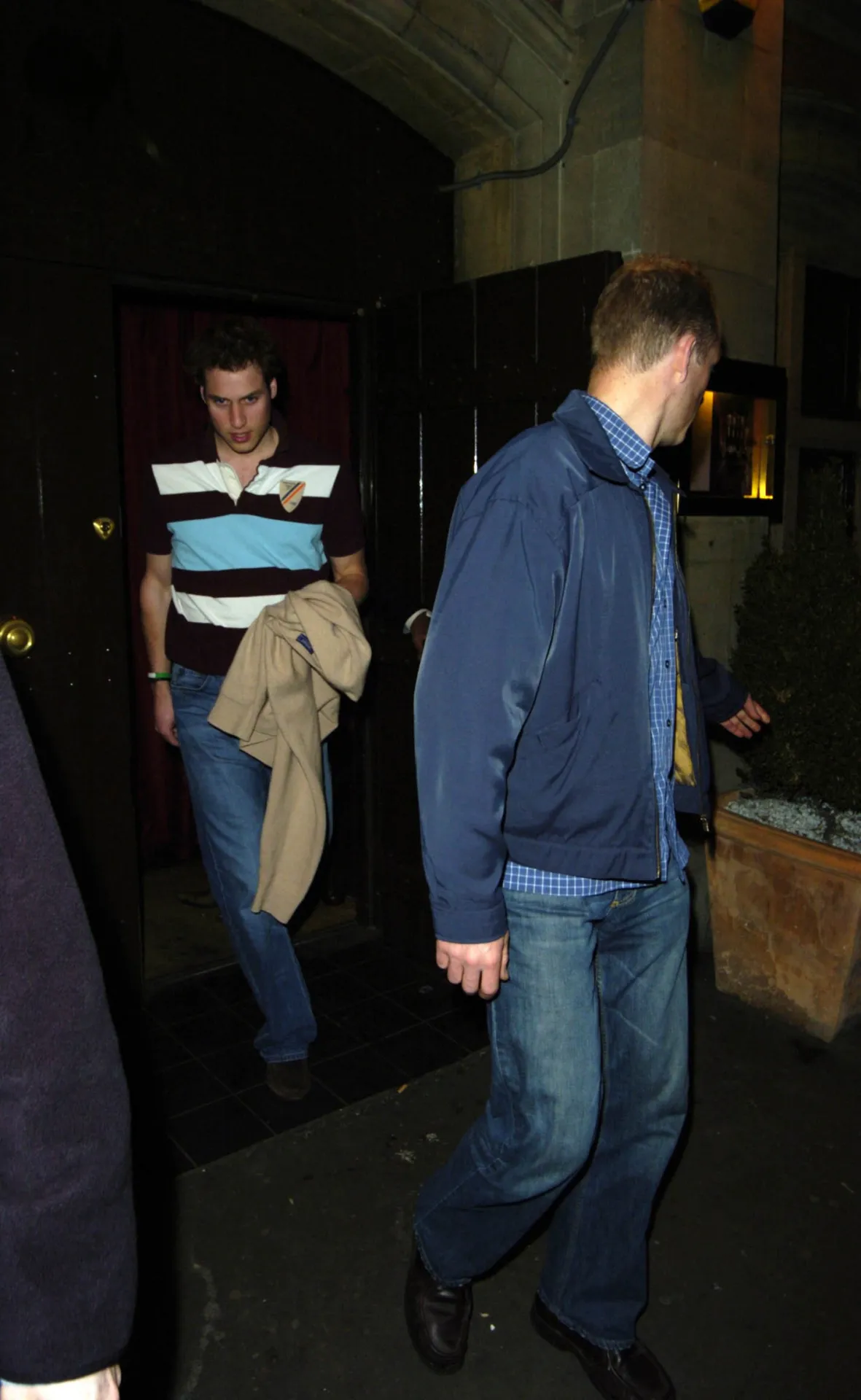 Prince William in a striped rugby shirt with a jacket over his arm, coming down from a doorway at night, with a man in a blue