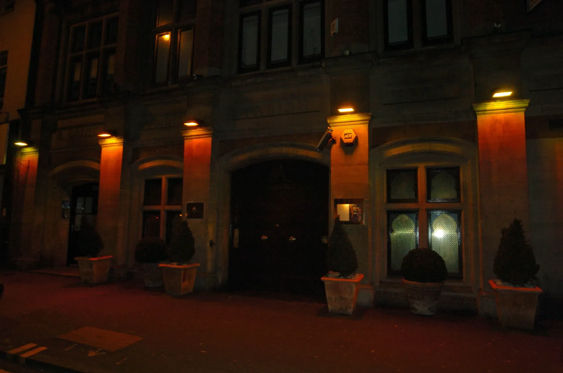 The stone front of the Sugar Hut bar and restaurant at night, lit by orange wall lamps, with potted shrubs by the arched door